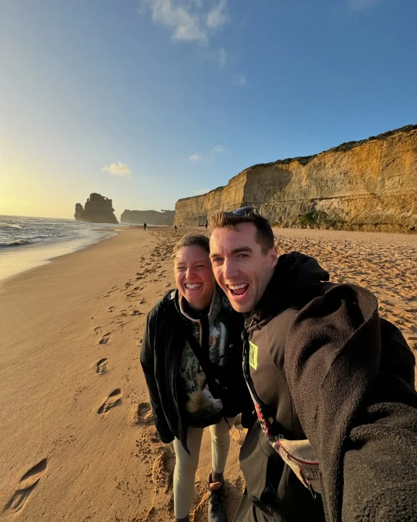 Glen and Mado smiling on the beach during their Great Ocean Road trip in Australia, with stunning cliffs and ocean views in the background