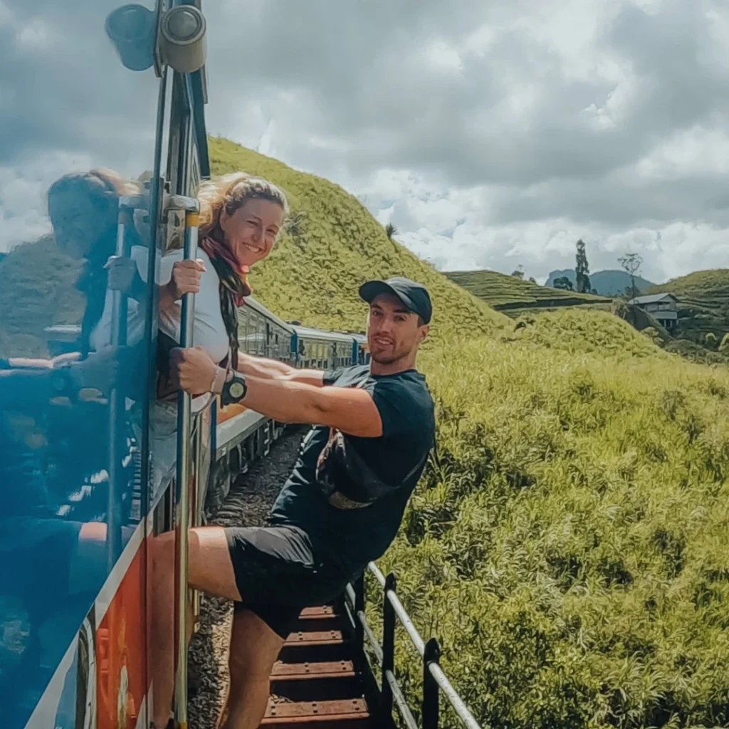 Glen and Mado hanging from a moving train in Sri Lanka, enjoying the scenic green landscape.